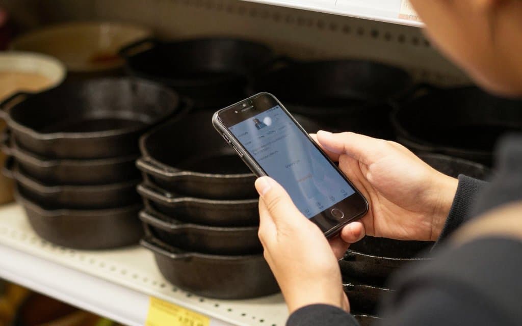 Shopper checking cast iron skillet value on a phone beside pans on a thrift shelf
