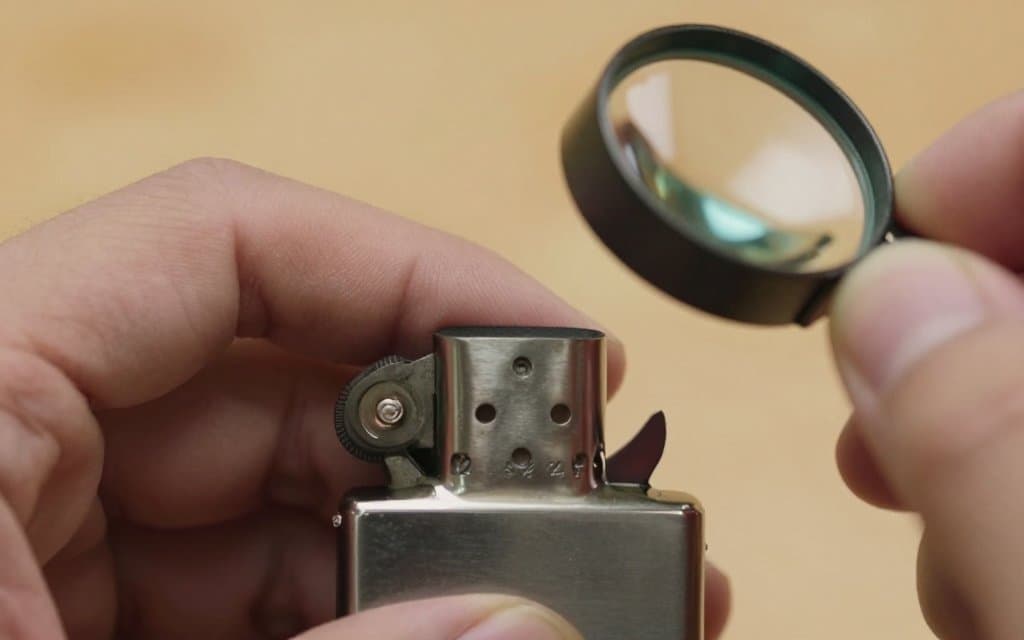 A hand using a loupe to inspect a Zippo lighter bottom stamp for a fake date code.