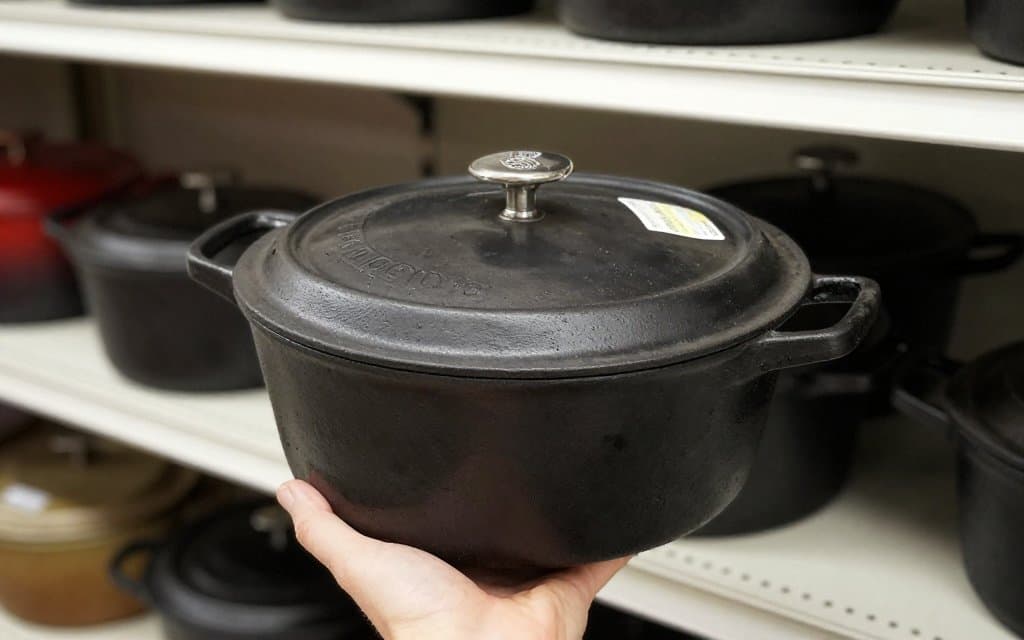 Shopper lifting a stained vintage Le Creuset Dutch oven off a thrift store shelf to check its weight